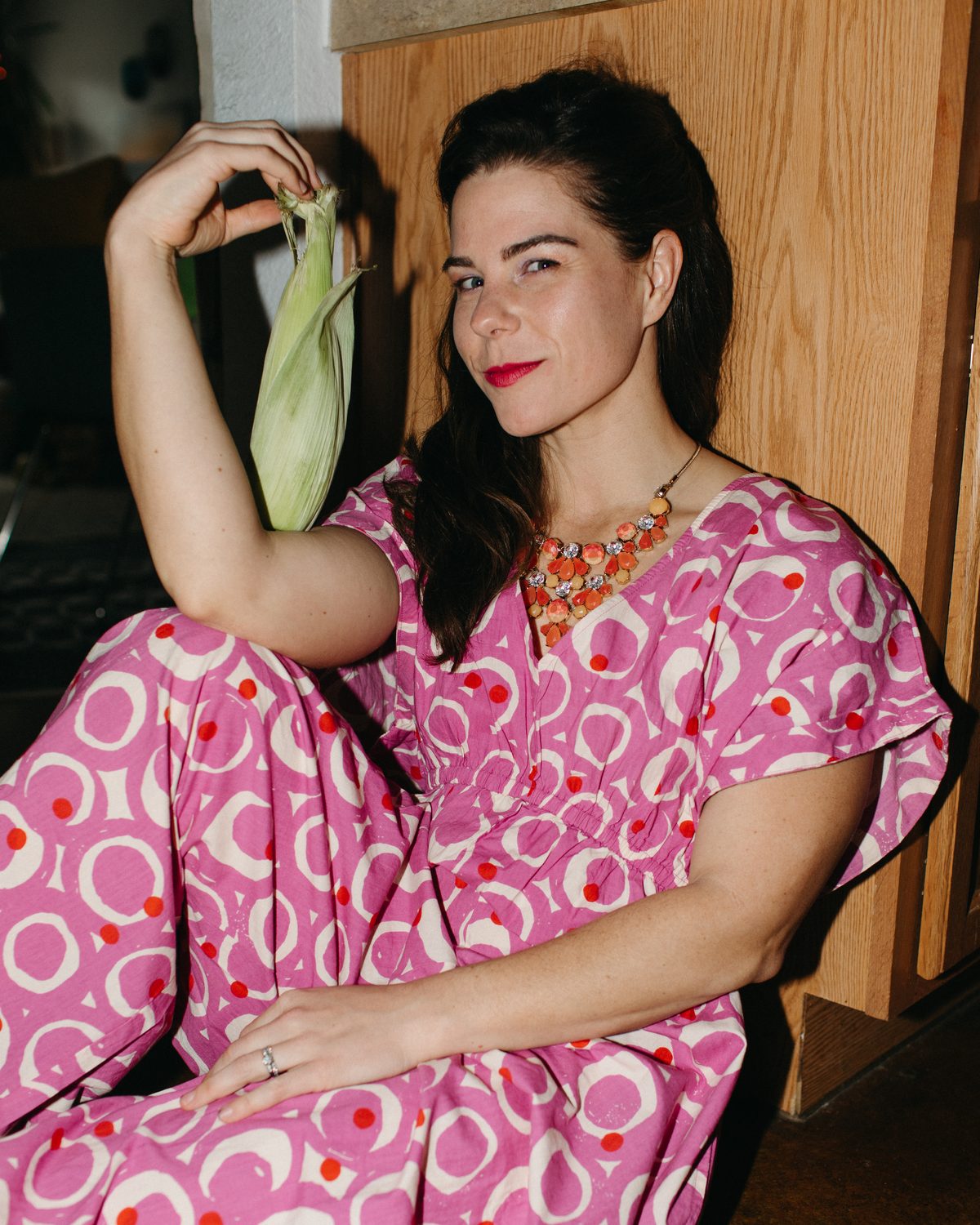 Jessi Roesch seated in a pink patterned dress, holding a husk of corn