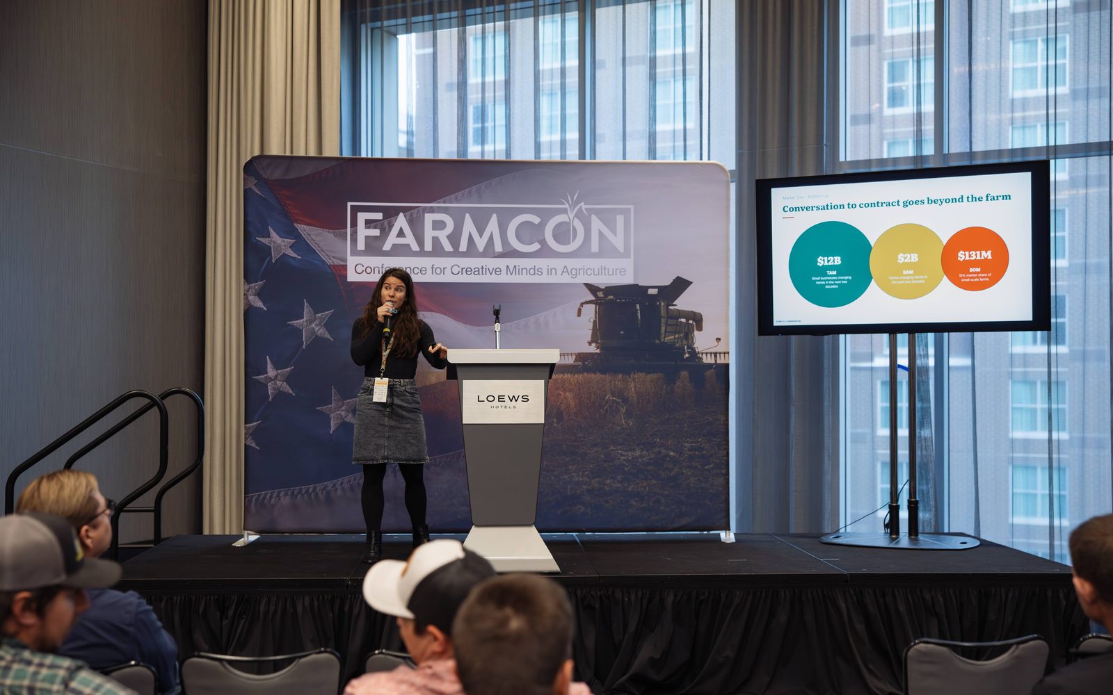Jessi presenting from a podium at FARMCON with a combine on the backdrop